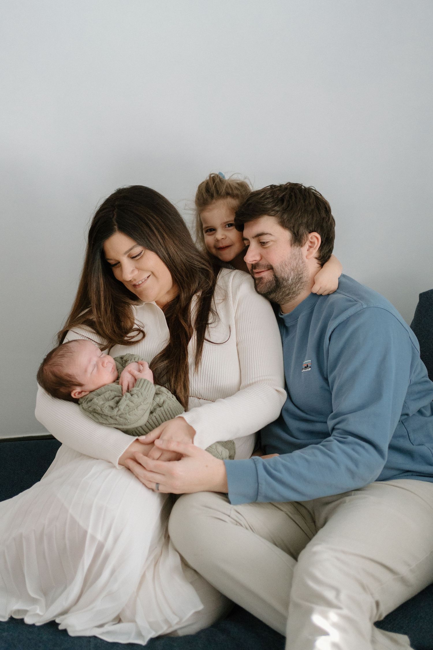 A smiling family of four sits together, mother holding newborn while toddler hugs father from behind.
