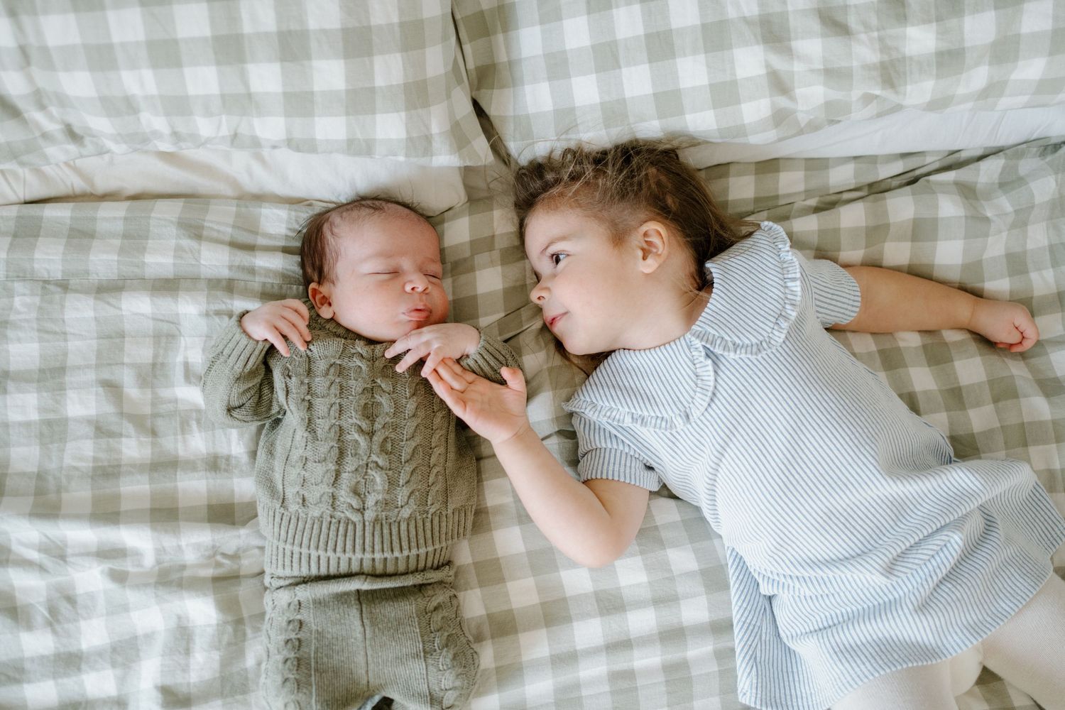 A newborn baby and toddler sibling lie together on a plaid bed, the older child gazing lovingly.