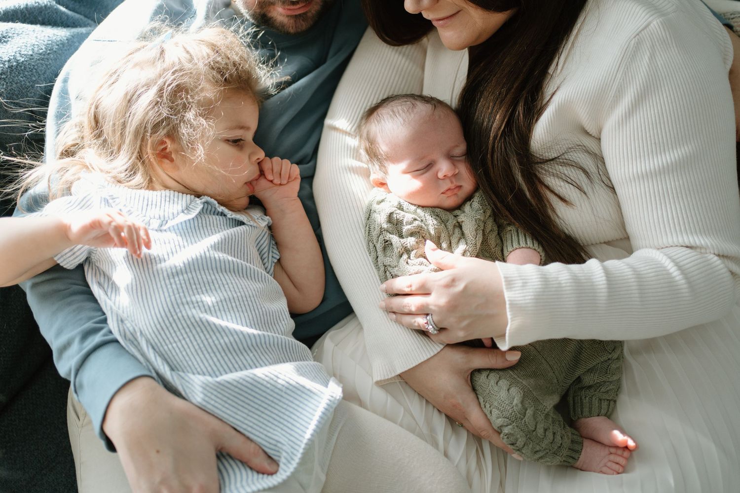 A family of four sits together, parents cradling a newborn while a toddler leans in close.