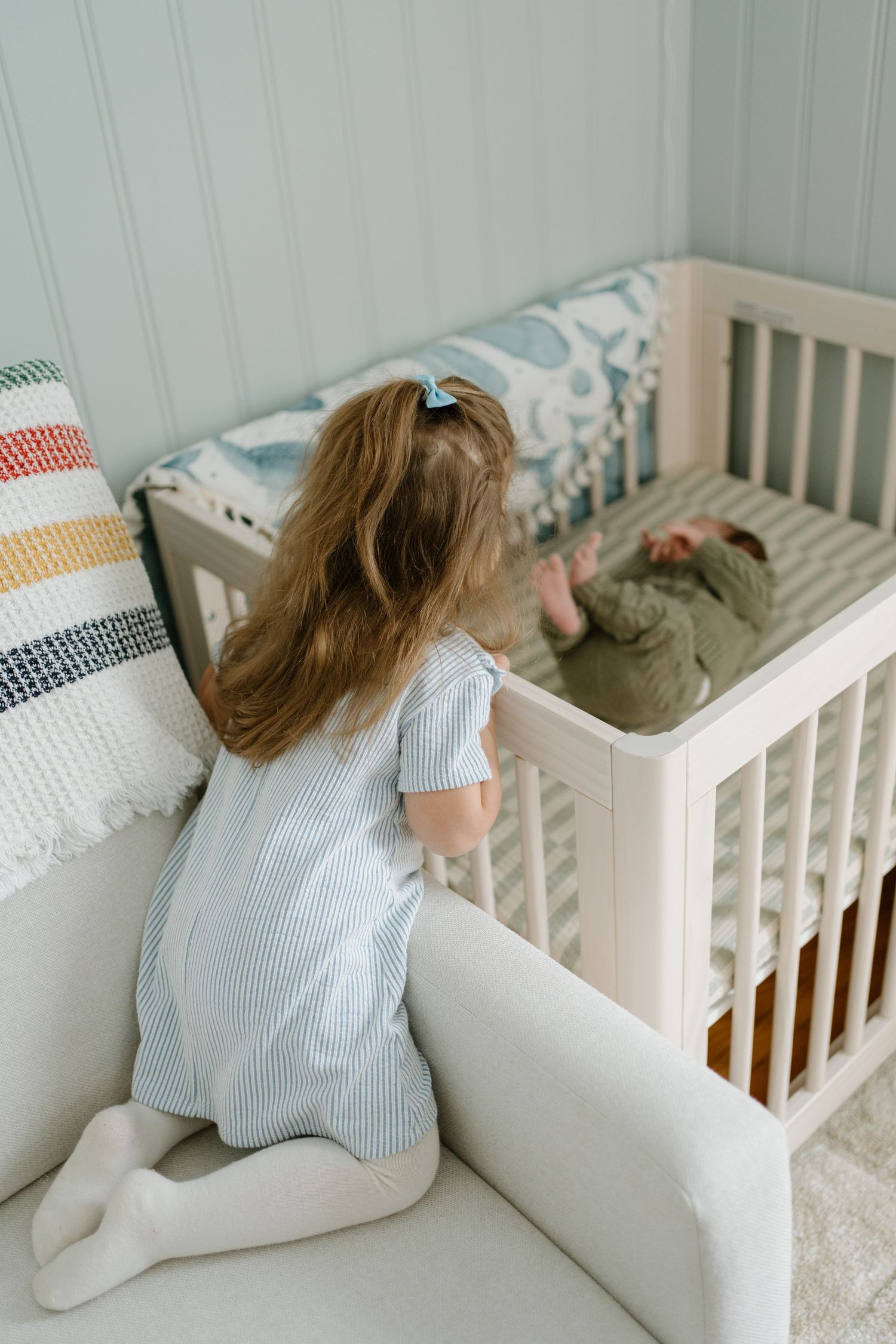 Toddler girl in striped dress kneeling on a sofa looking at a newborn baby lying in a white crib in a soft nursery.