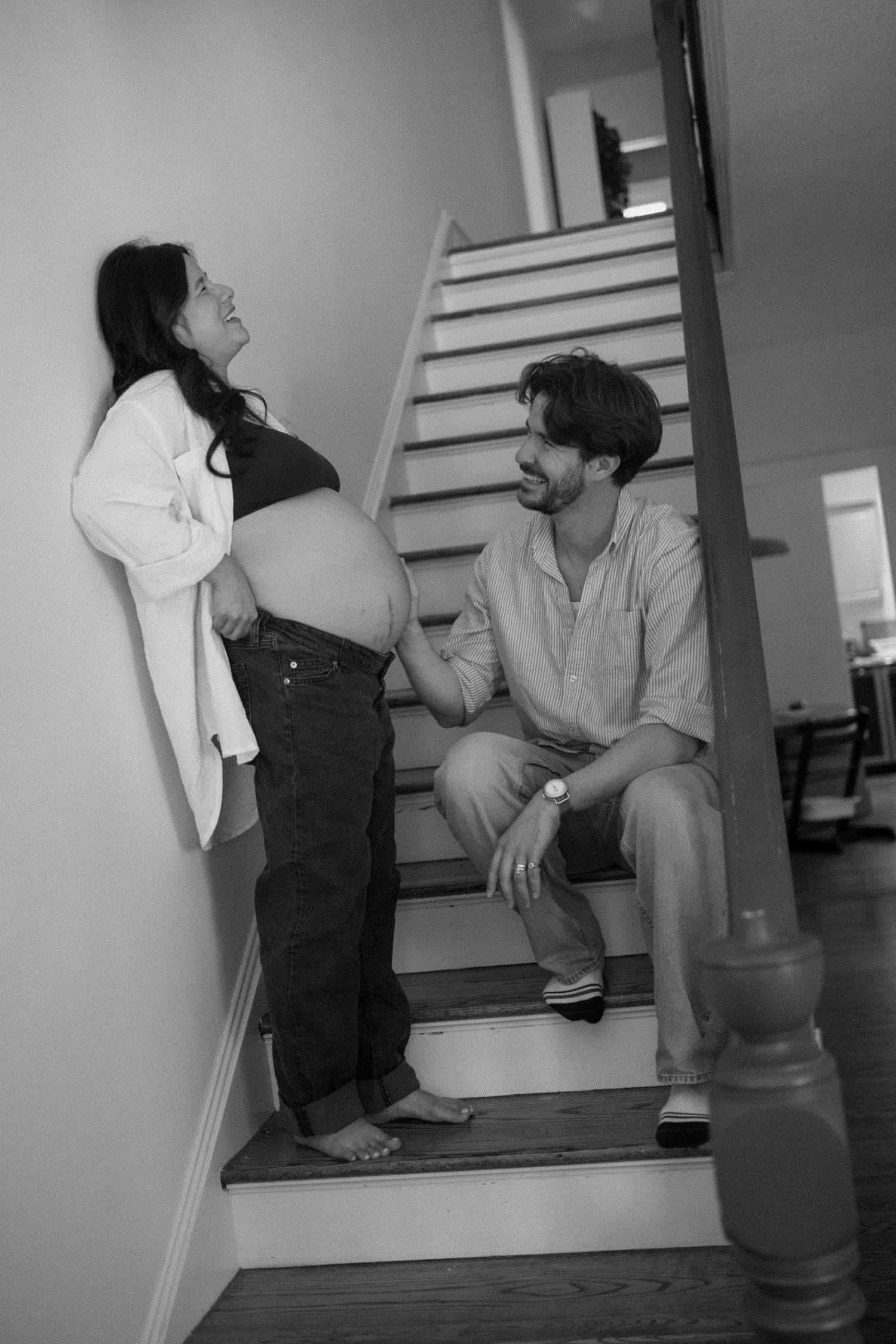 Black and white photo of a pregnant woman standing by stairs while her partner sits on steps smiling at her baby bump.