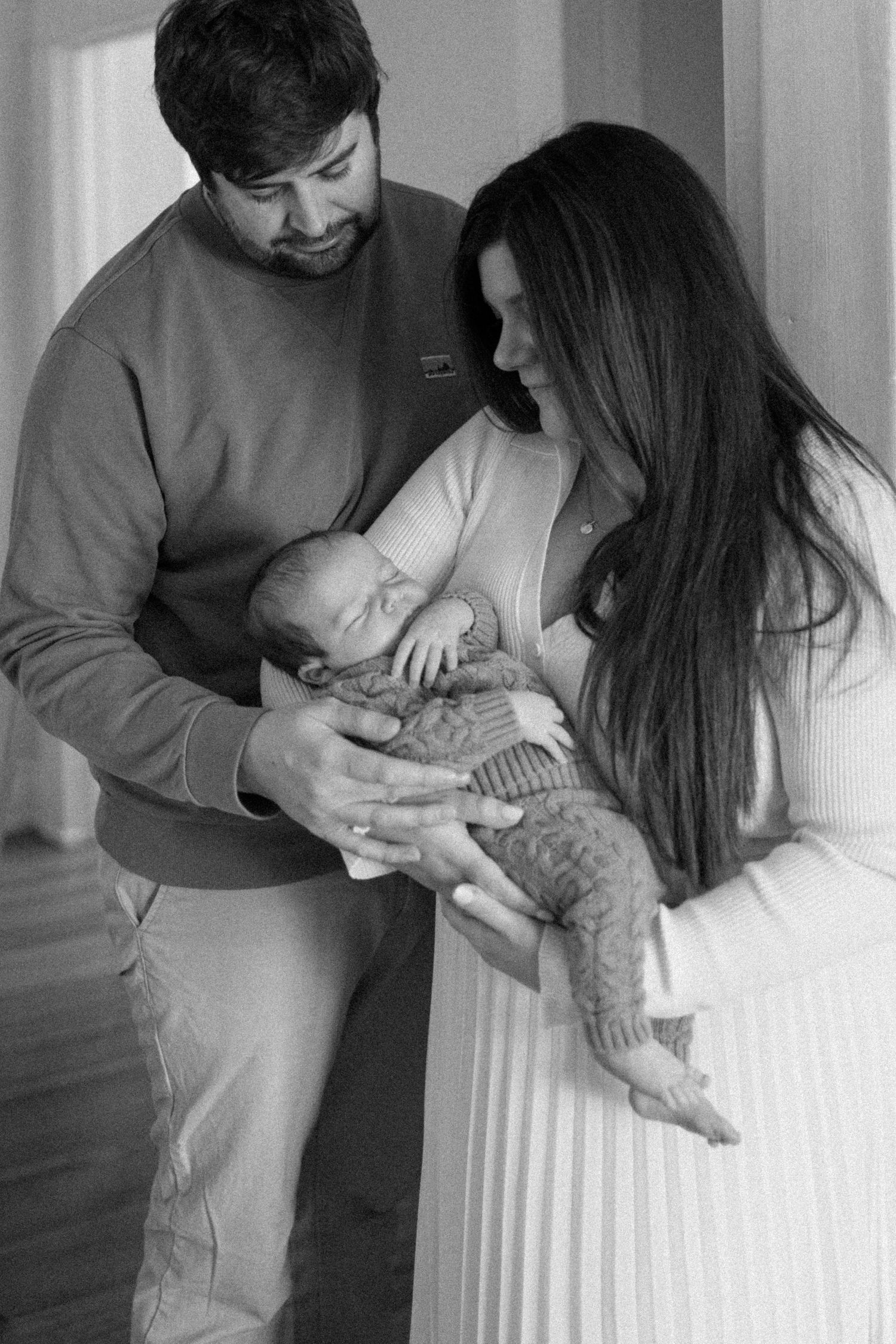 A couple gazes lovingly at their newborn baby held between them, captured in black and white.