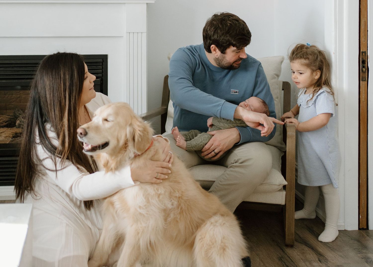 Family with golden retriever, newborn, and toddler relaxing by fireplace in cozy home.