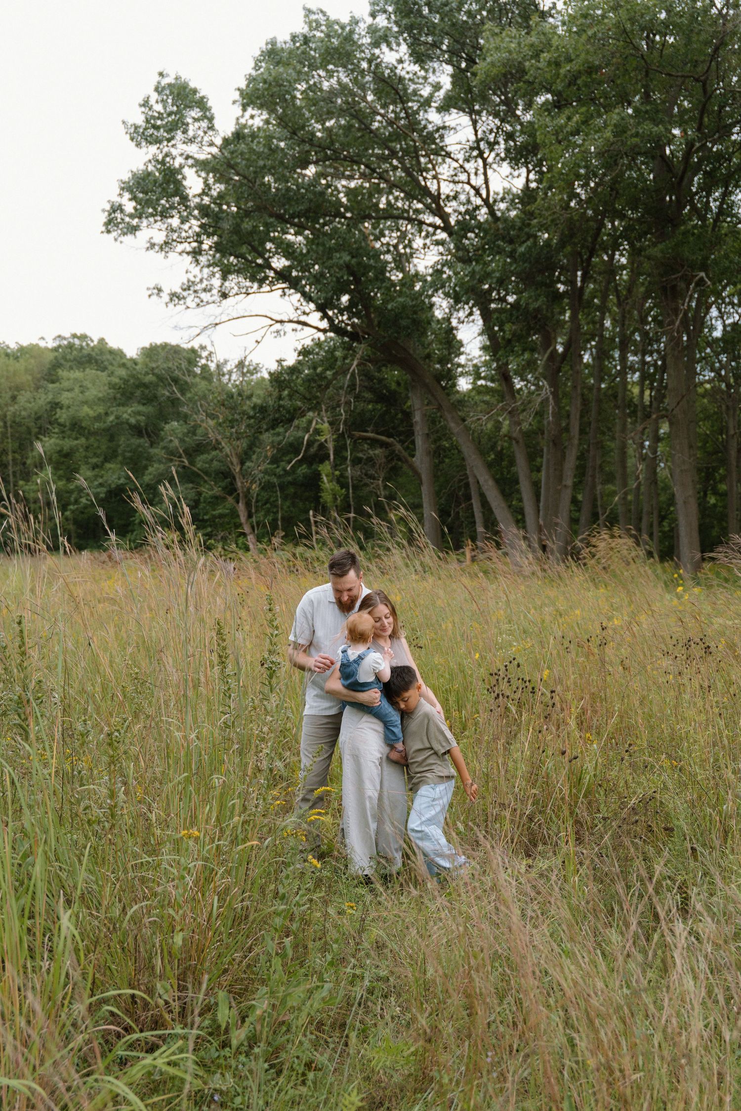 Family of four embracing in tall golden grass field with lush green trees in background during outdoor photo session.