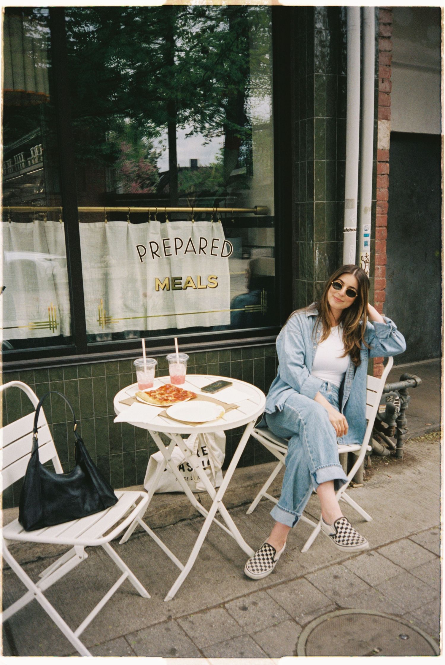 Woman in denim jacket and checkered shoes sits at outdoor cafe table in front of Prepared Meals shop window.