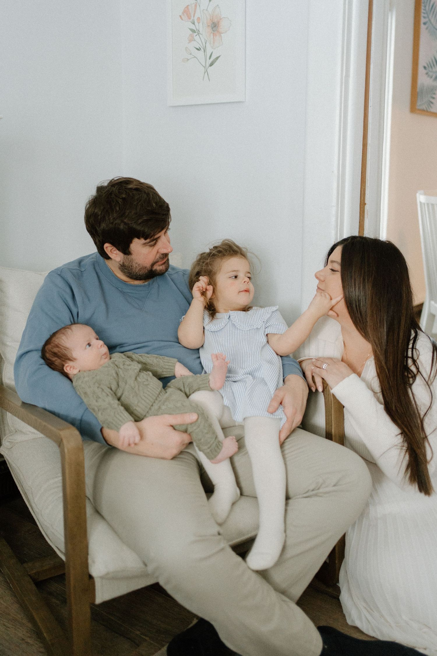 Family of four sitting in chair, dad holding newborn while toddler touches moms nose.