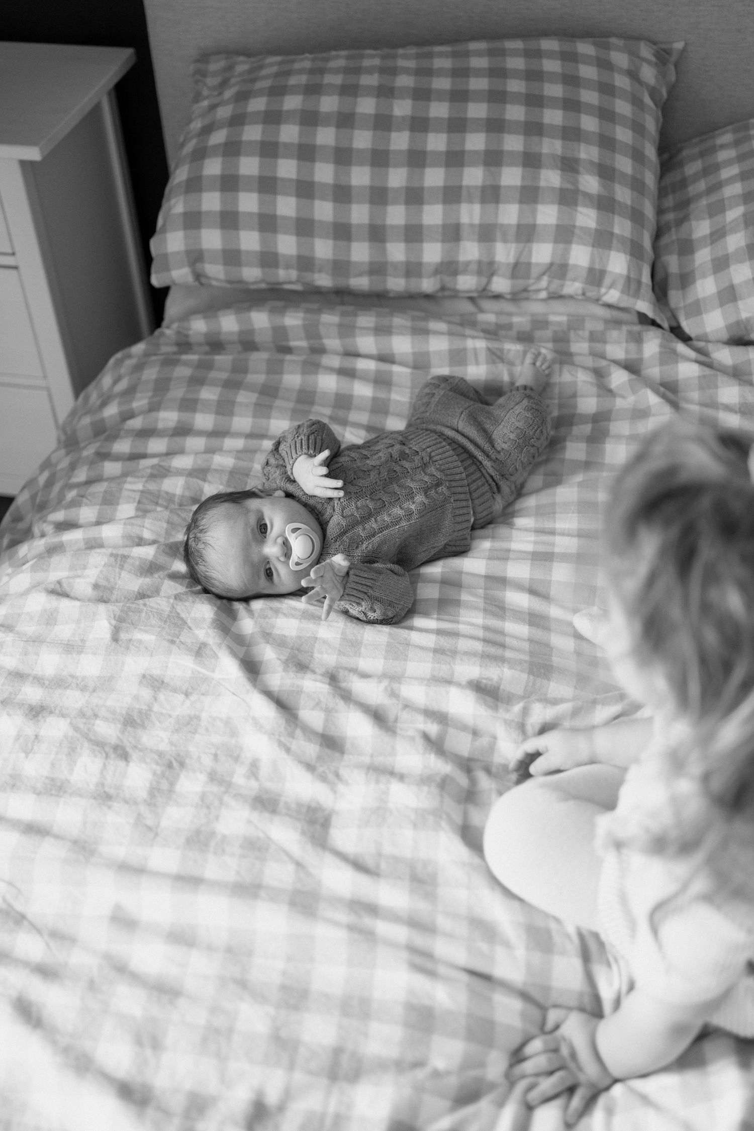 Black and white overhead photo of newborn baby lying on plaid bedding while older sibling sits nearby on the bed.