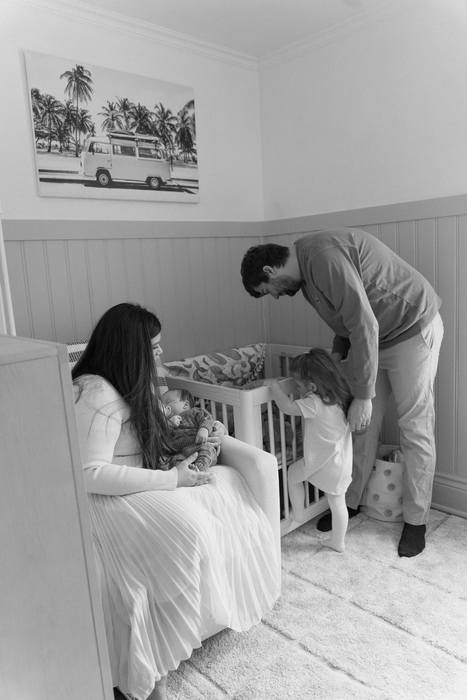 Black and white photo of parents and toddler in nursery, mother holding newborn while father helps young child at the crib.