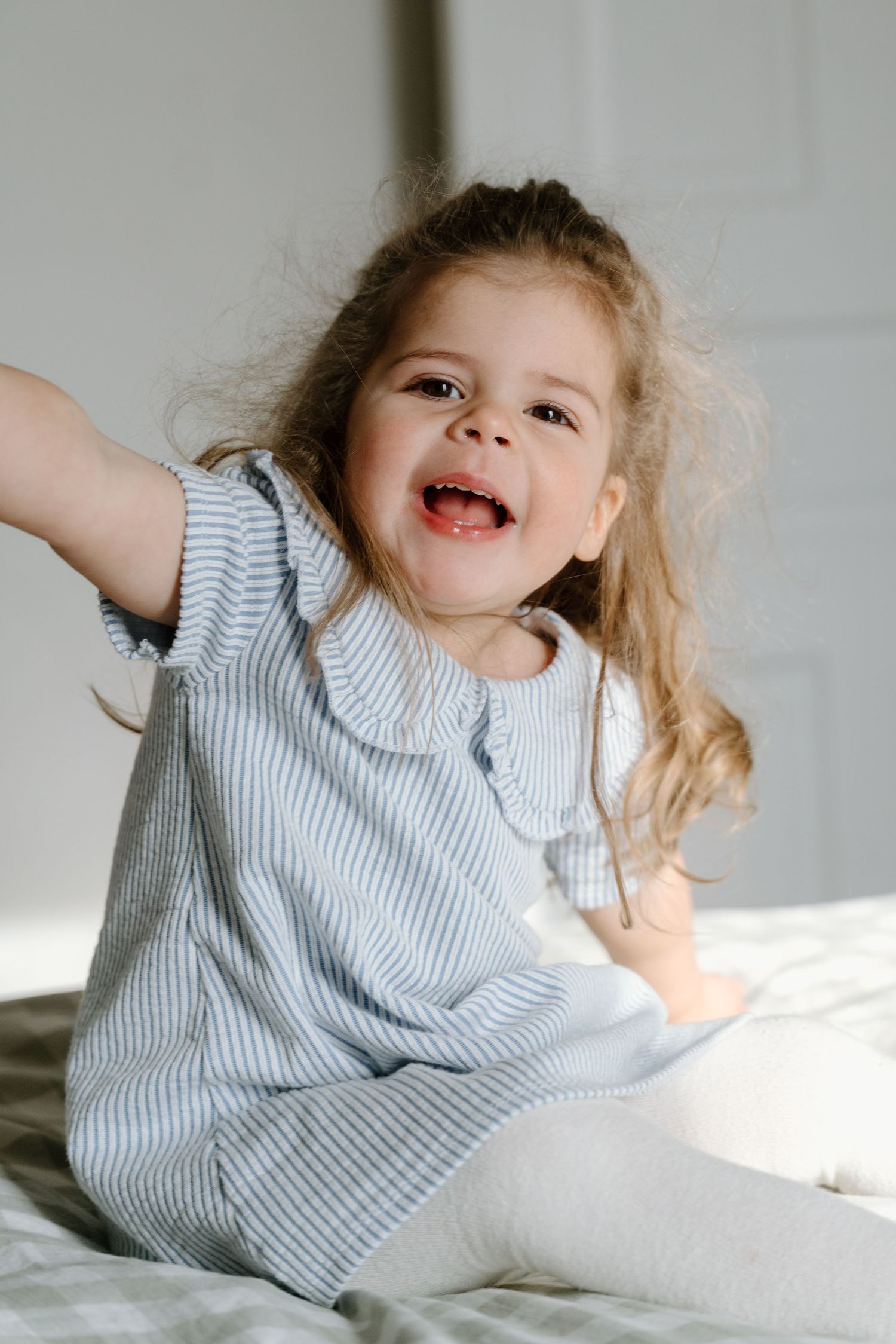 Adorable curly-haired little girl in a striped dress laughing and playing on a bed.