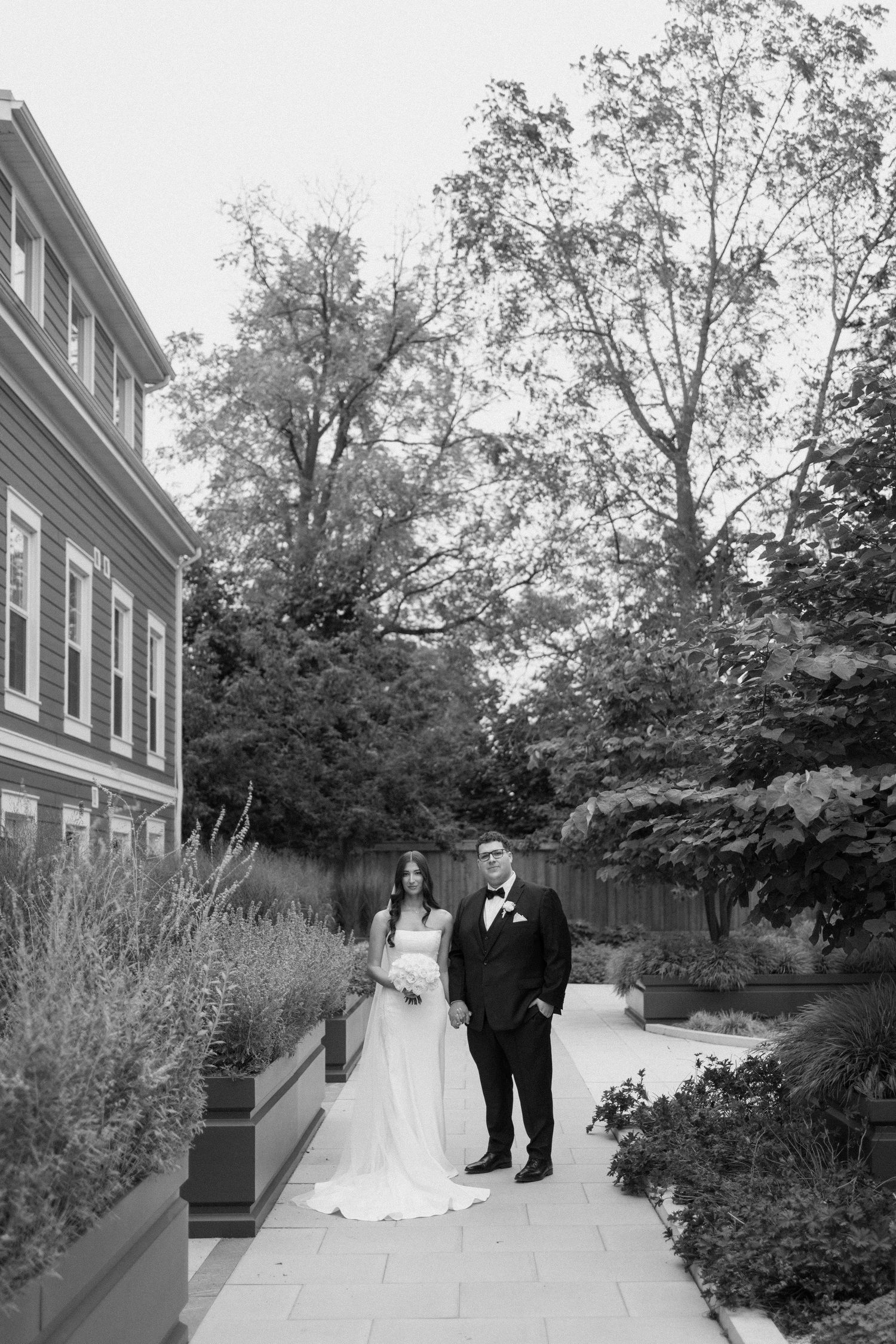 Black and white photo of bride and groom posing on garden path beside elegant building surrounded by lush greenery.
