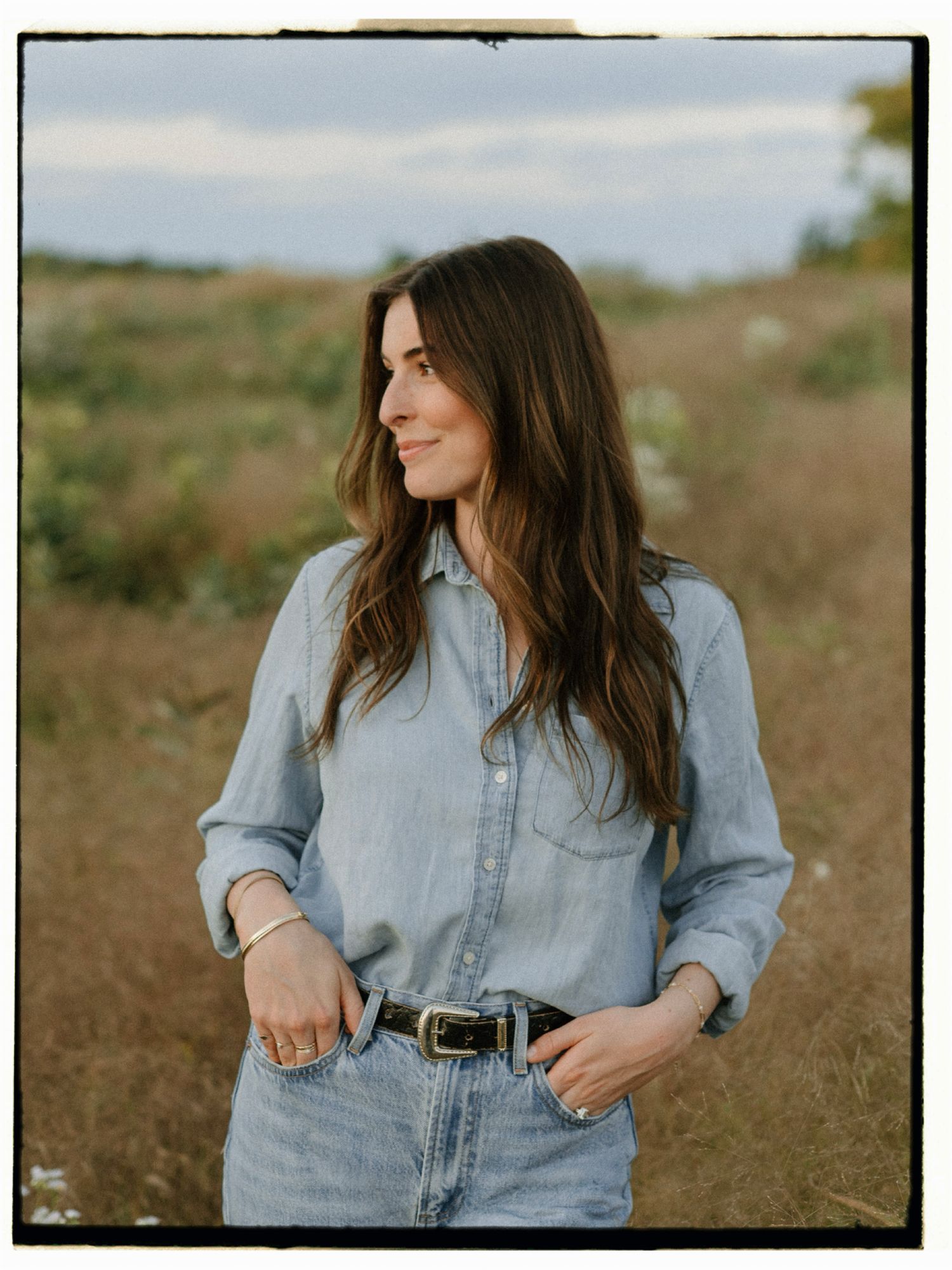 Woman in light blue denim shirt and jeans with black belt standing in a field on a cloudy day.