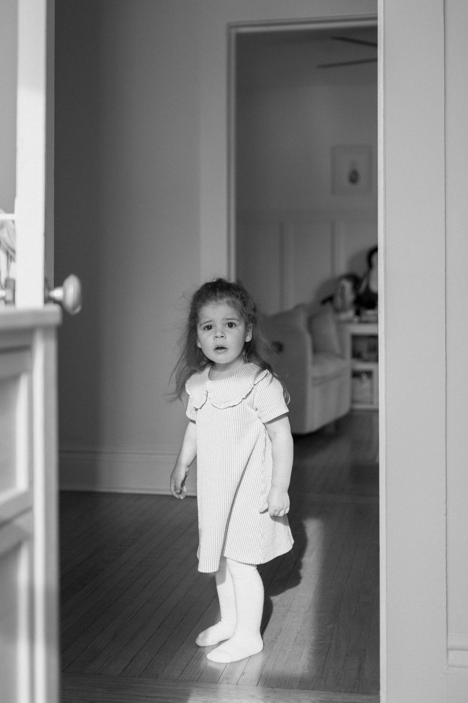 A young girl in a white dress and tights stands in a doorway of a home, captured in black and white.