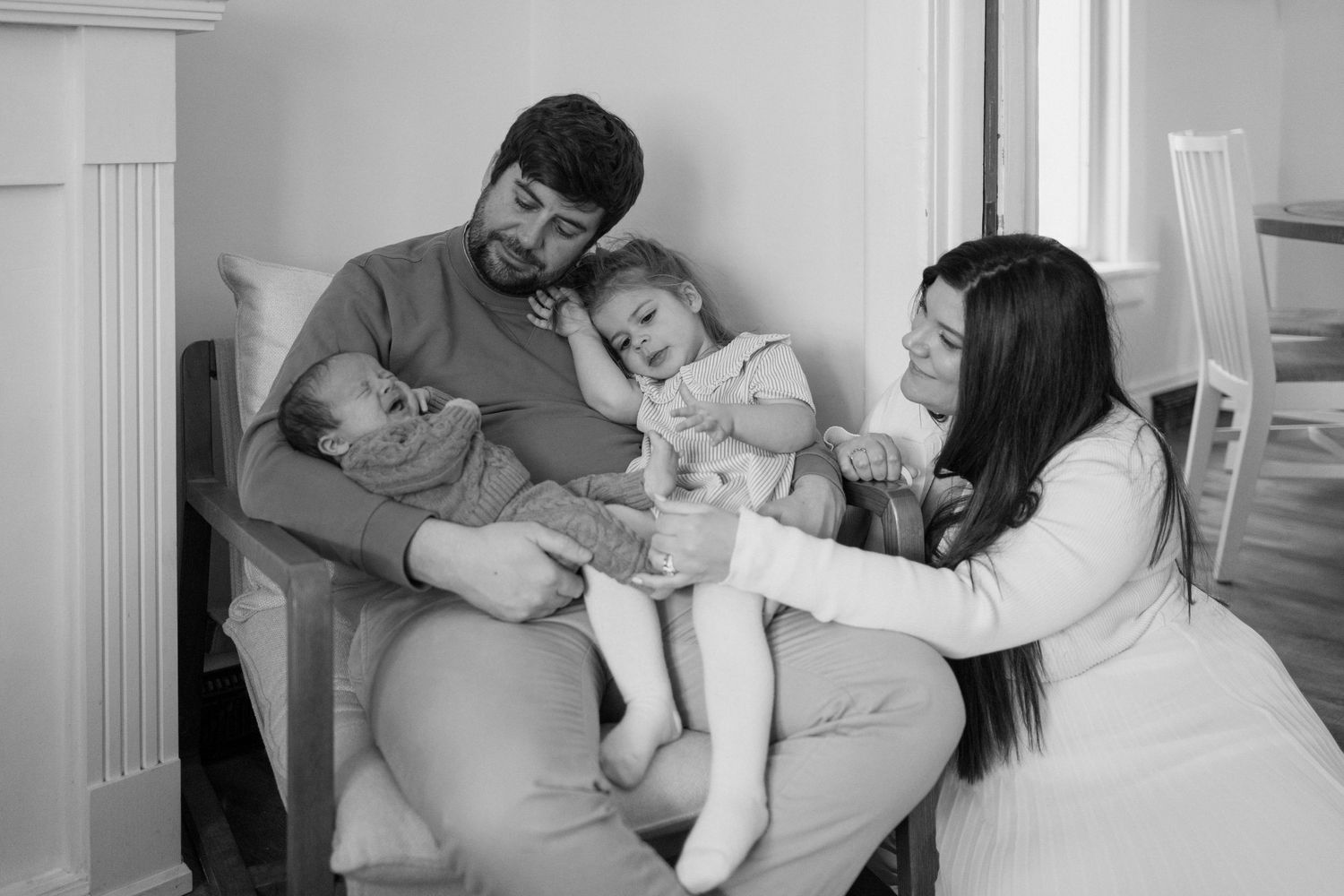 Black and white photo of parents with newborn and sleepy toddler in cozy indoor setting.