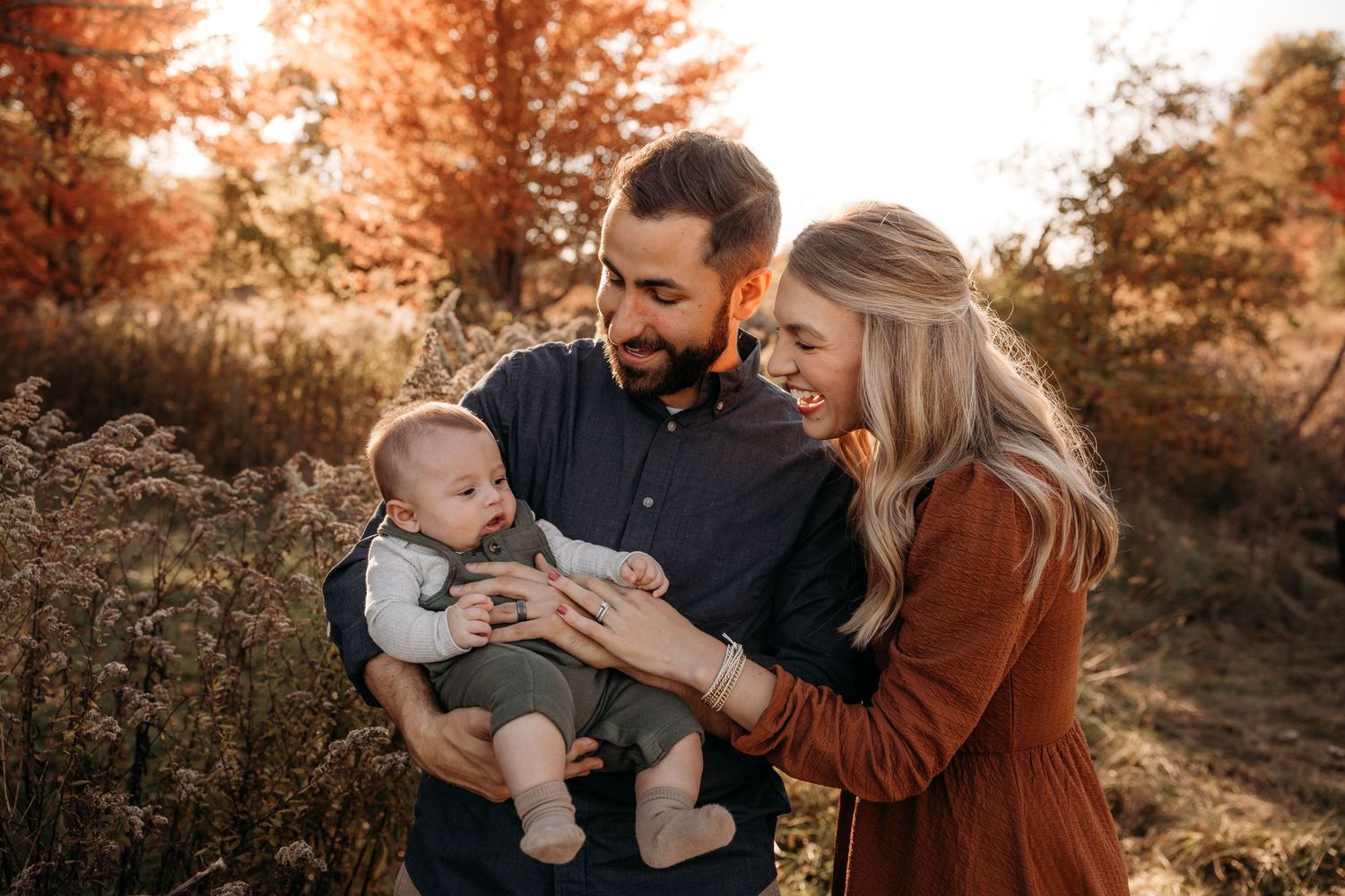 Young parents smiling at their baby boy during a fall photo session with vibrant orange autumn trees in Branson, MO.