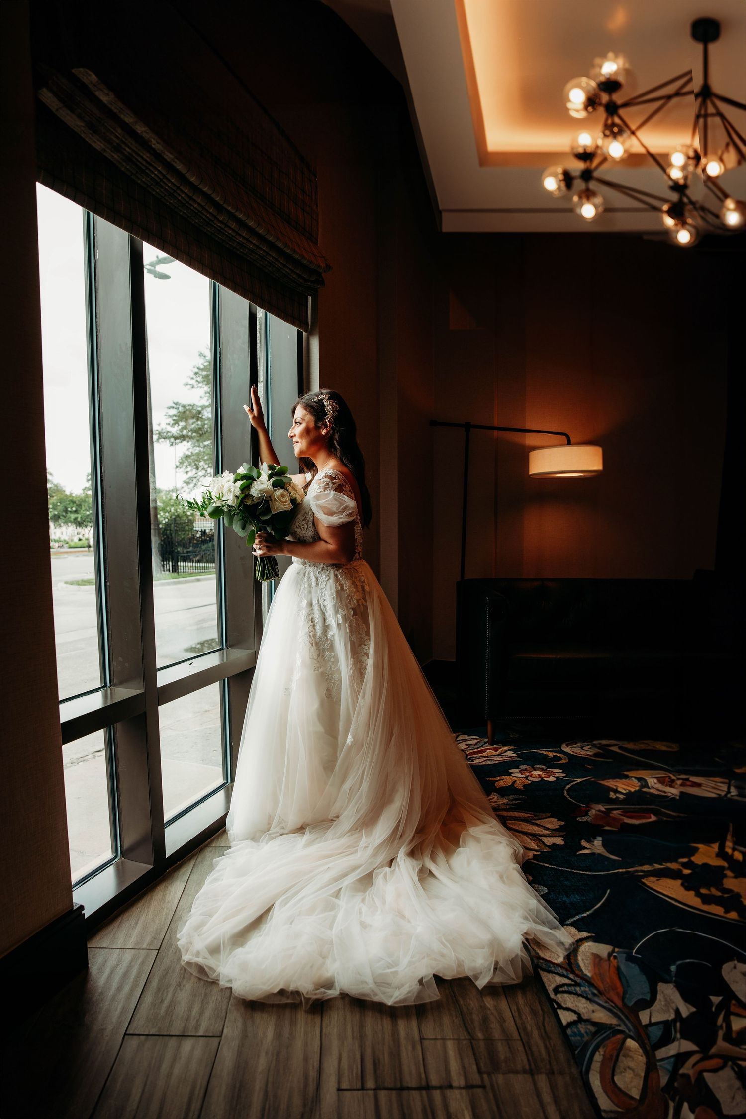 Elegant bride in a lace wedding gown holding a white bouquet while looking out a large window at a Branson, MO wedding venue.