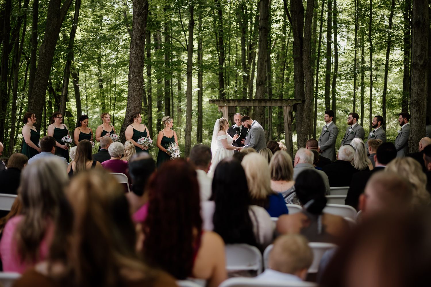 Outdoor wedding ceremony in a wooded forest setting with white chairs and guests seated beneath tall trees.