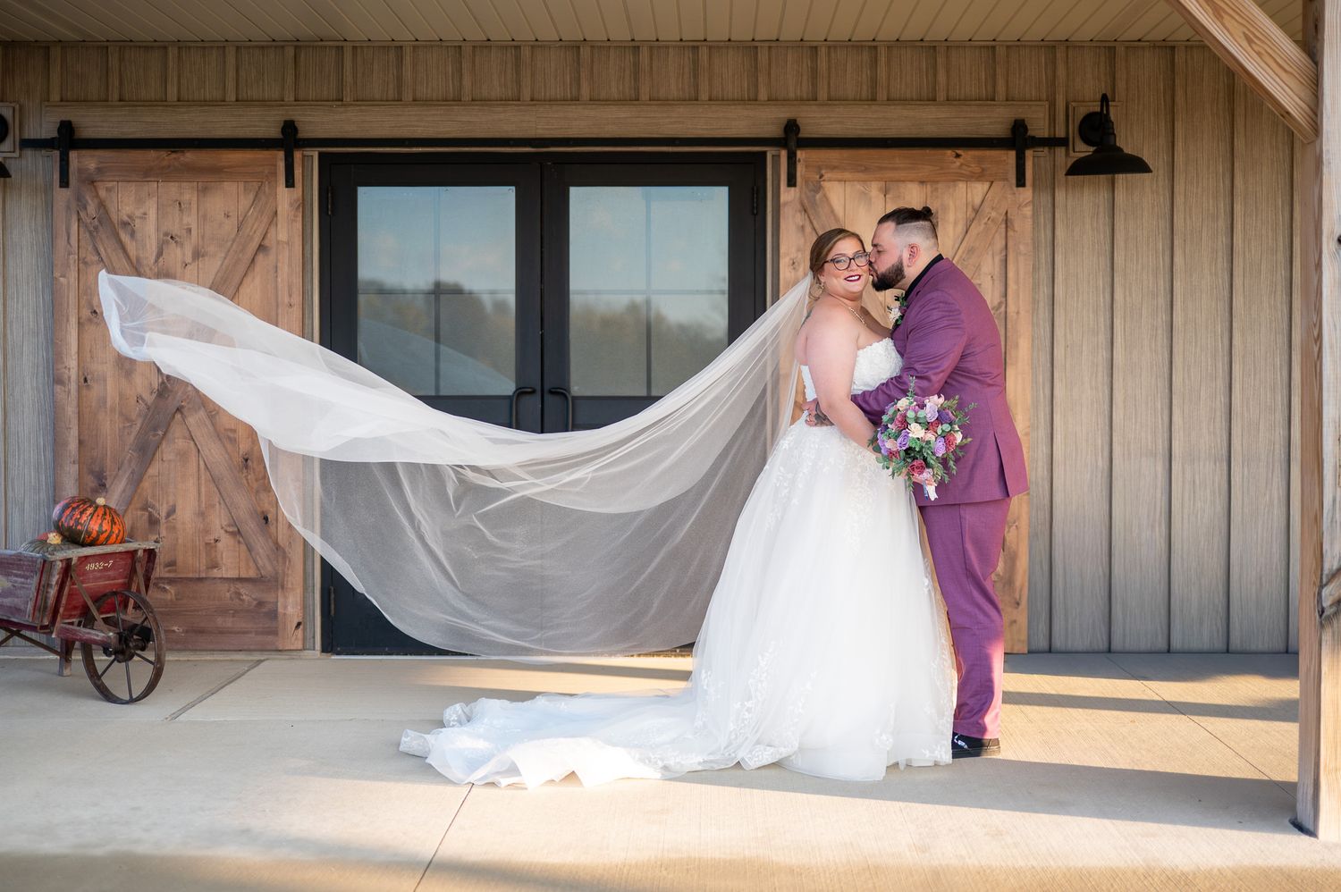 A flowing white wedding dress veil catches the wind in a rustic outdoor setting against wooden barn walls.