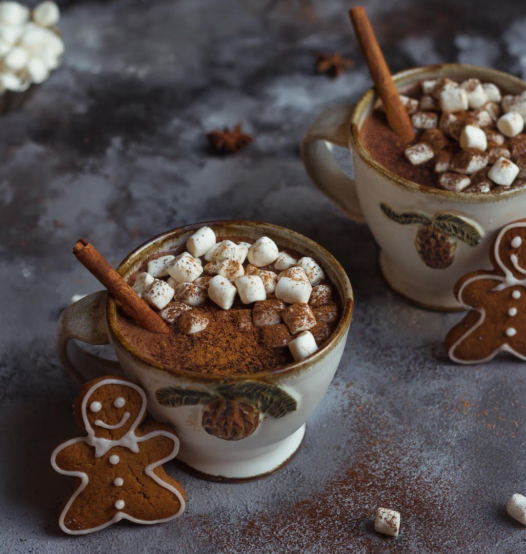 Hot chocolate topped with mini marshmallows and cinnamon sticks served with gingerbread cookies on dark background.