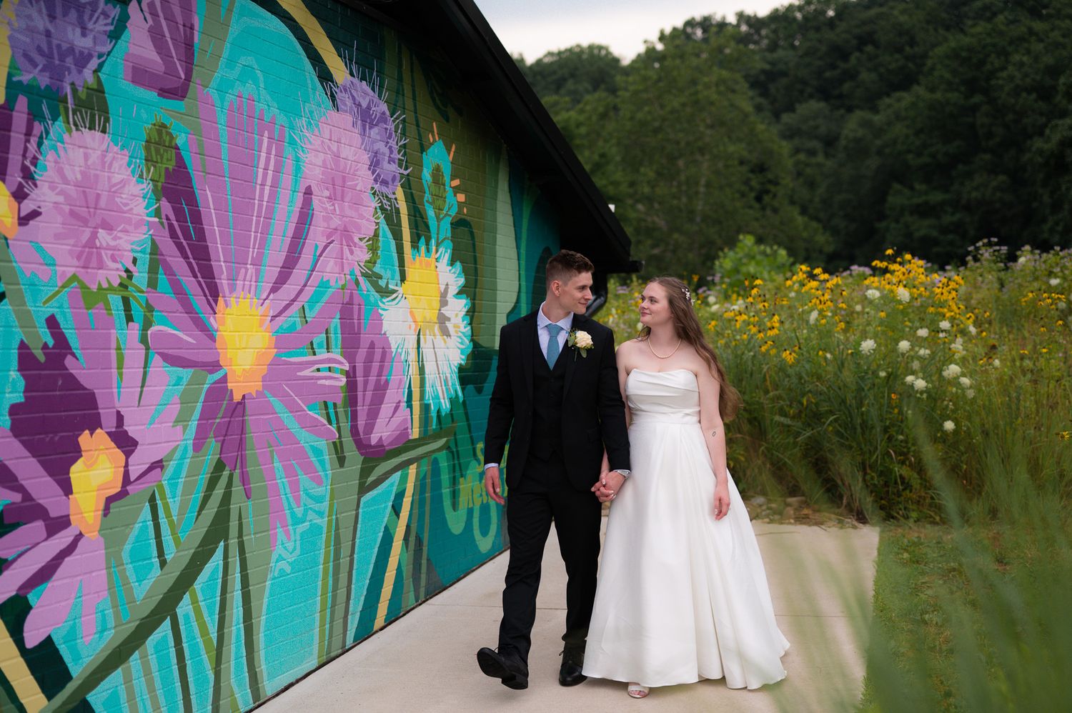 A colorful floral mural serves as a backdrop for a wedding photoshoot in a garden setting with wildflowers.