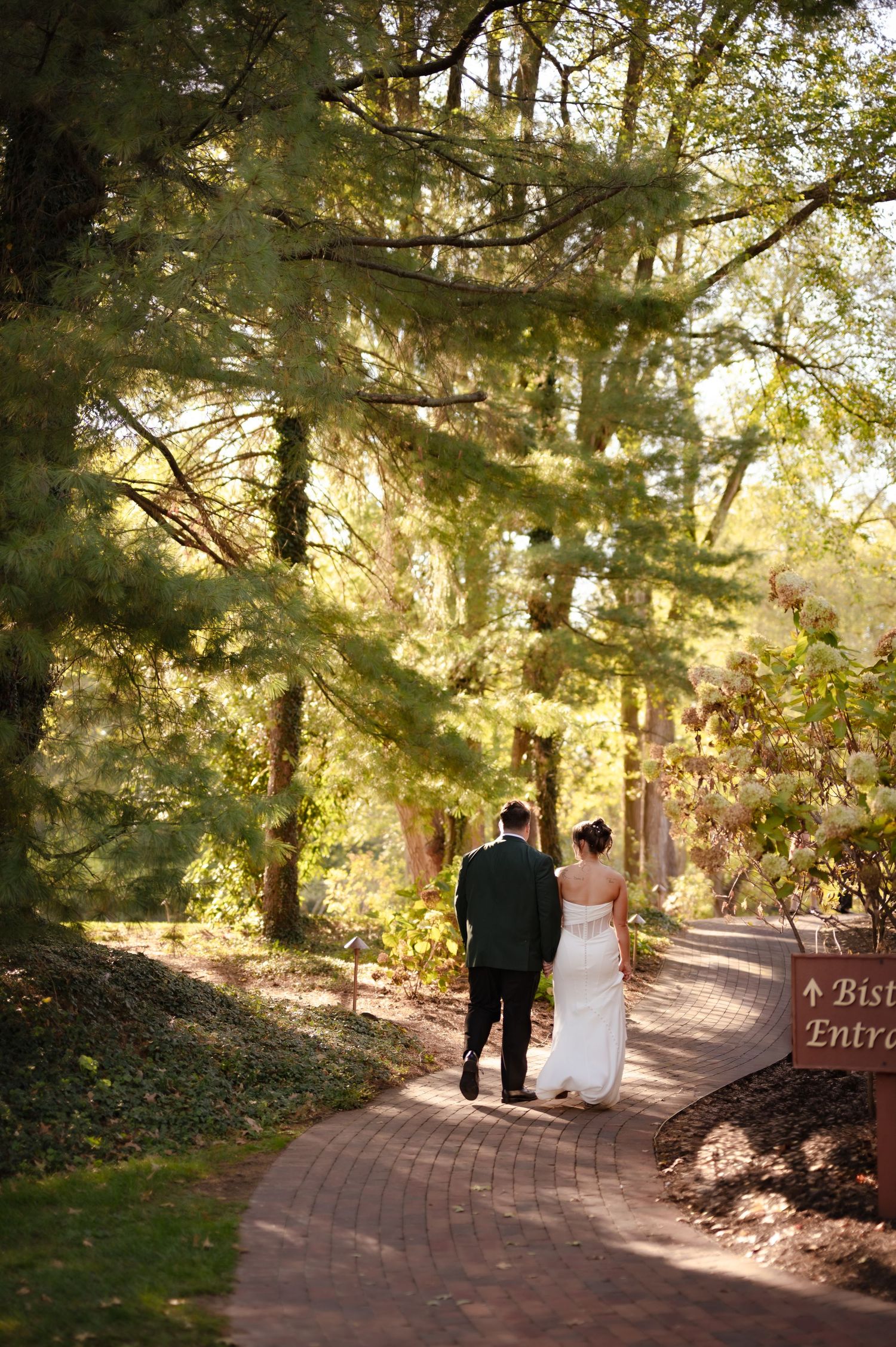 Couple walking down sunlit brick path surrounded by tall trees in romantic garden setting in Stark County, Ohio.
