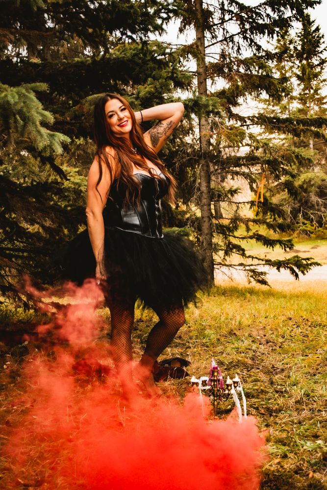 Gothic figure in black dress poses in forest with dramatic red smoke effect at sunset.