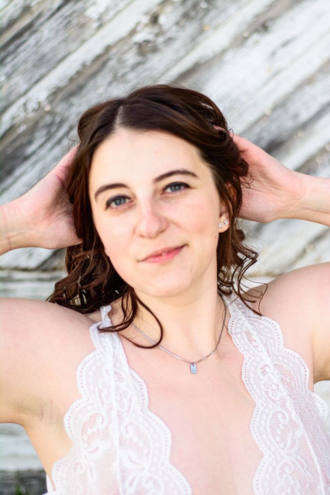 Portrait of person in white lace top with curly brown hair and delicate necklace against rustic background.