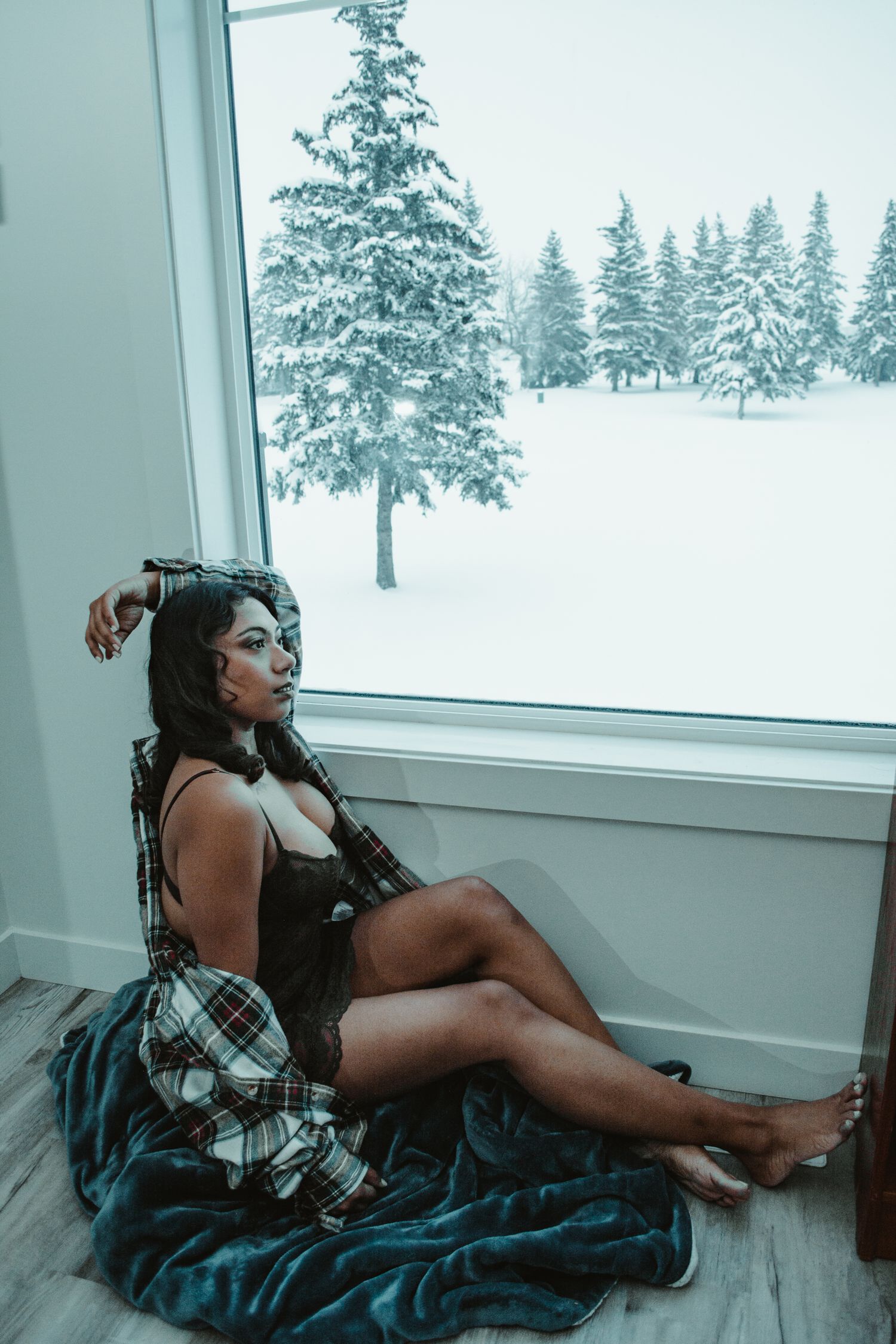 Model poses on hardwood floor by window overlooking snow-covered evergreen trees.
