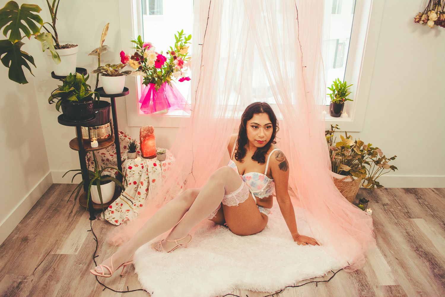 Romantic boudoir photo with pink tulle curtains, white rug, and floral arrangements in sunlit room.