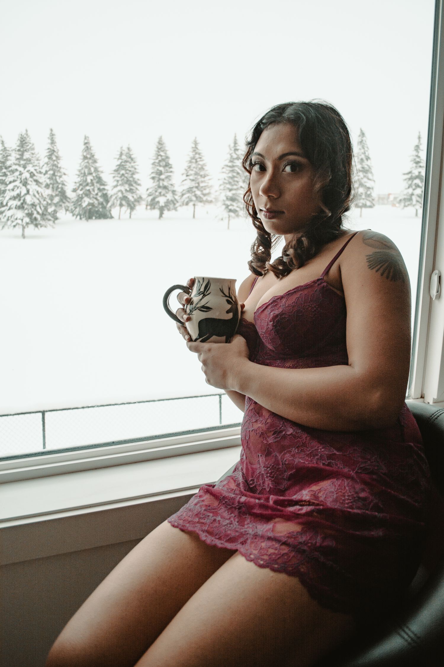 Woman seated by window holding ceramic mug with winter forest view.