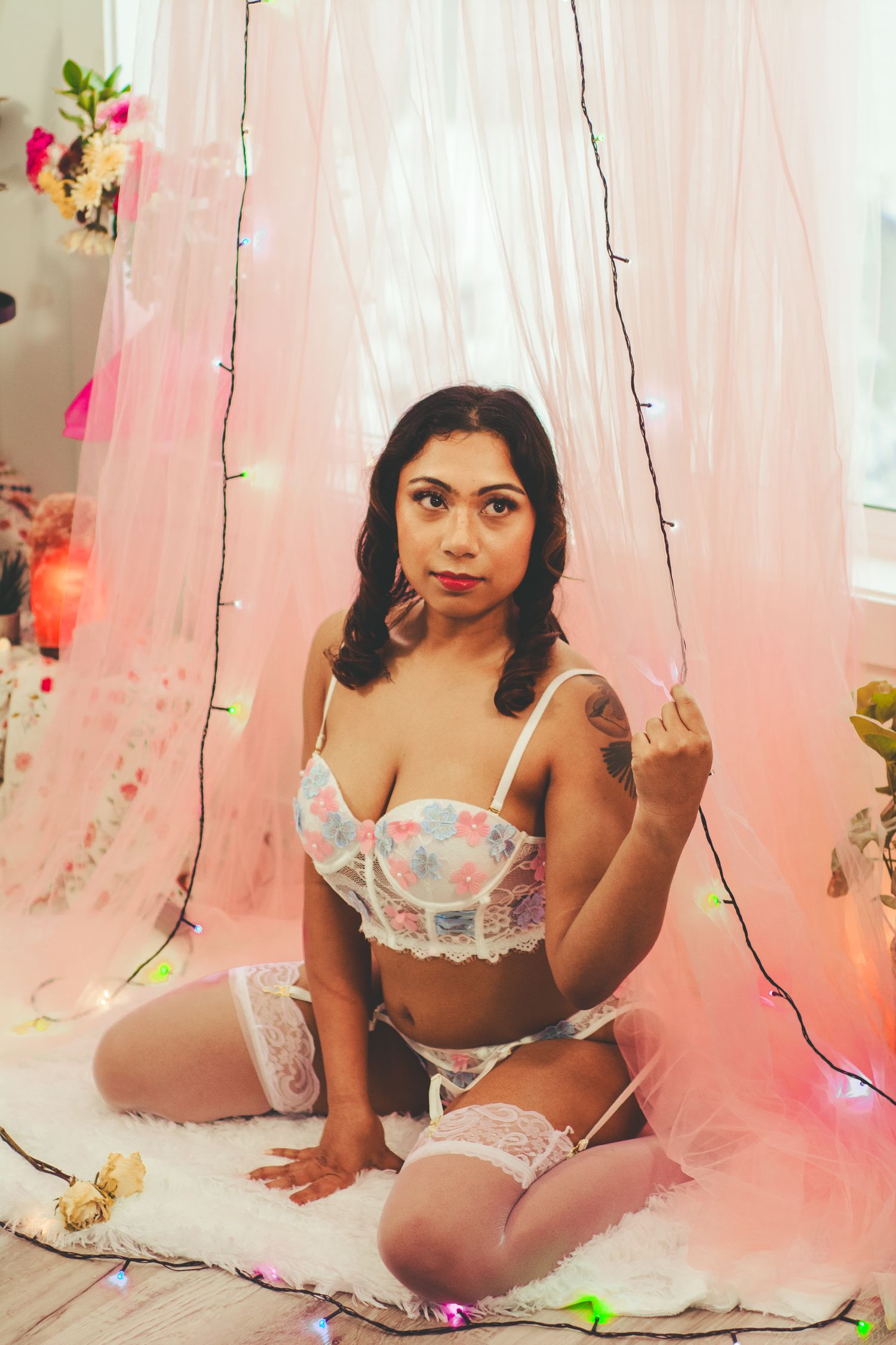 Romantic boudoir photo with pink tulle curtains, string lights and floral decorations in soft lighting.