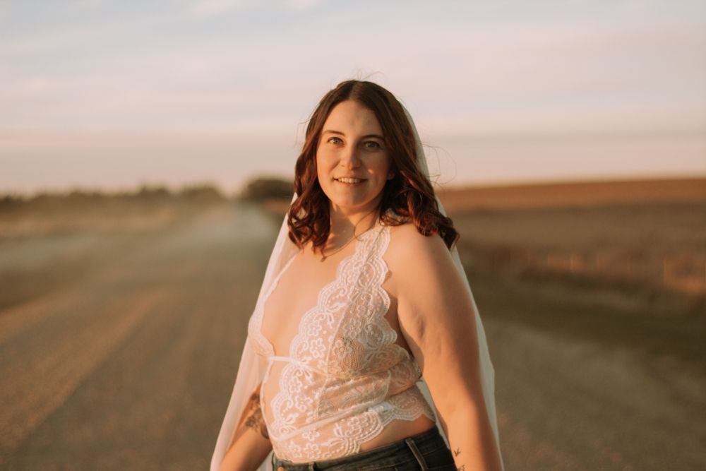 Portrait of person wearing white lace top standing on dirt road at sunset.
