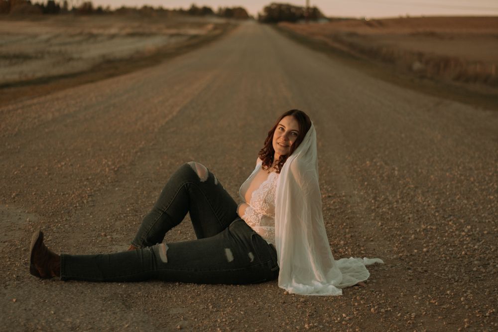 Person in white attire sits on empty country road during sunset.