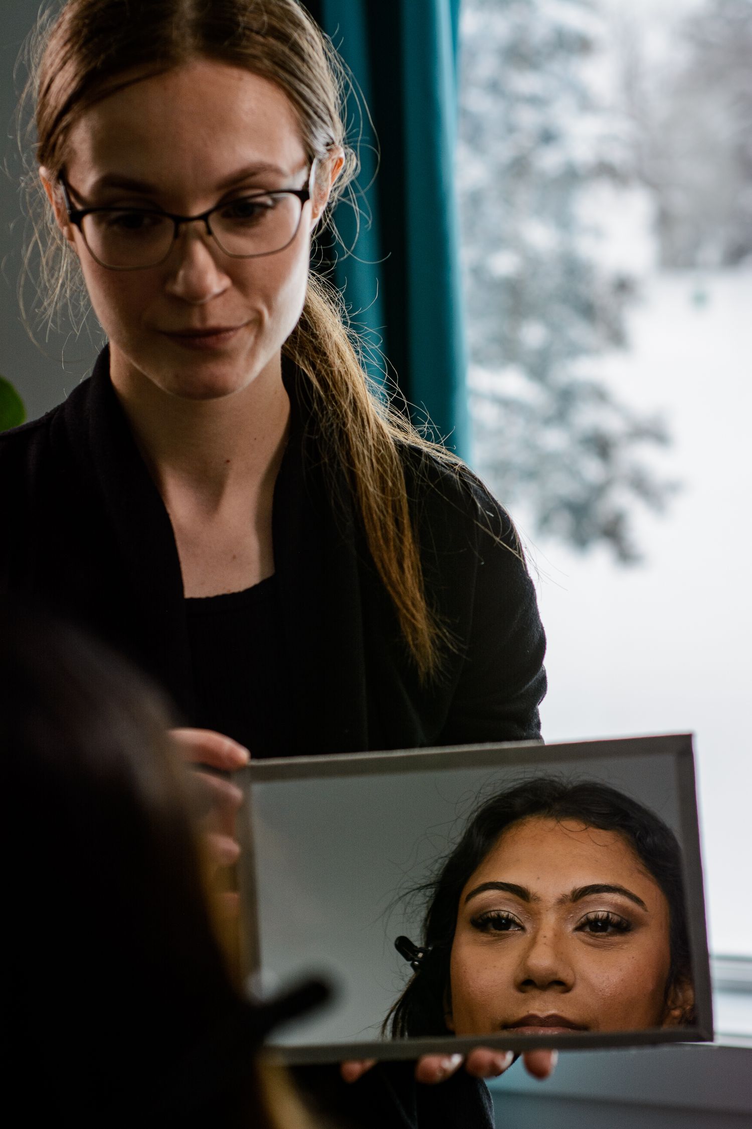 Professional makeup artist examines her work in a handheld mirror during a beauty session by a snowy window.
