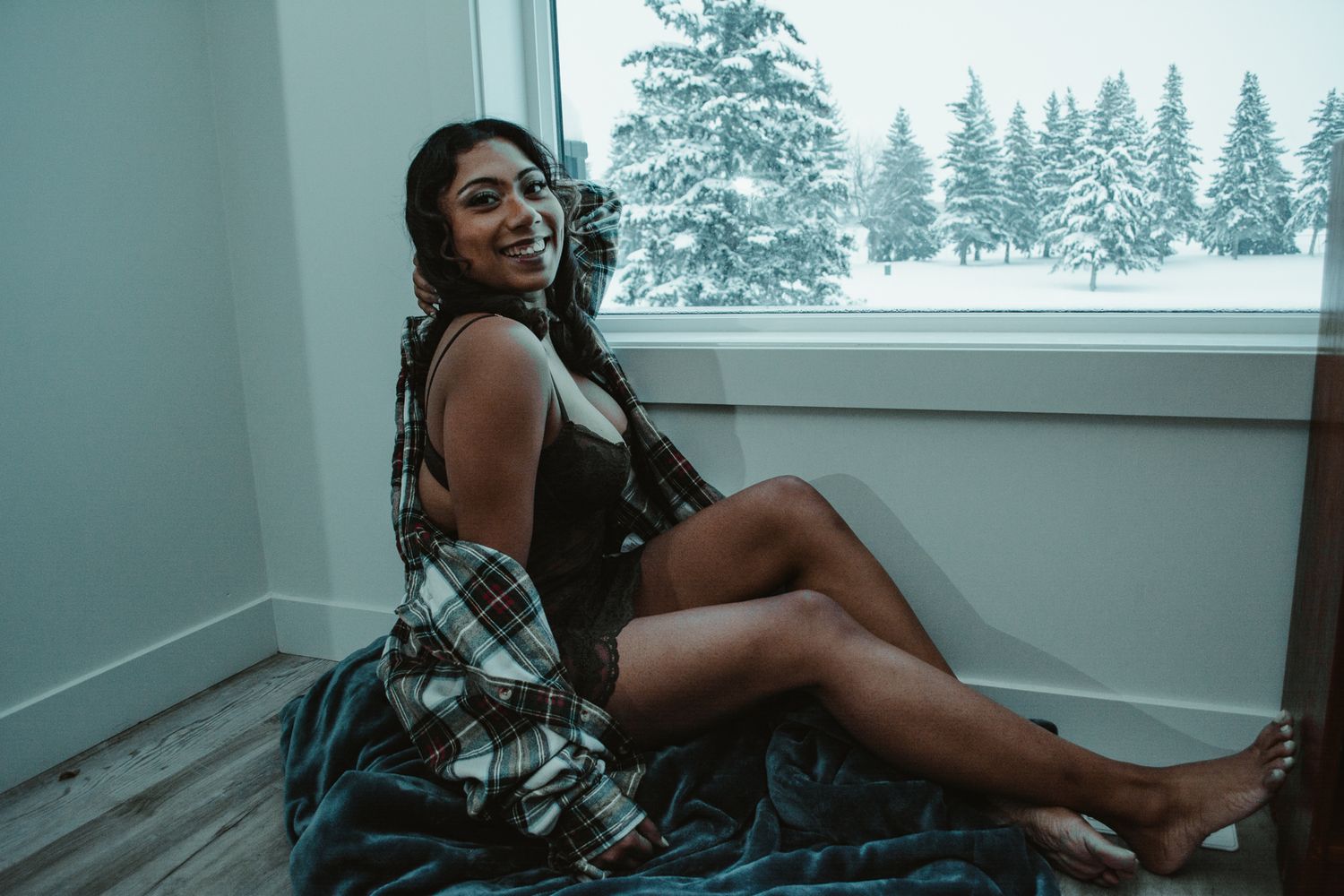 Boudoir photoshoot featuring woman in black lingerie against winter forest backdrop.