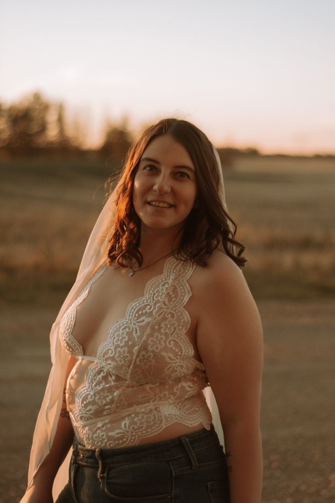 Woman in lace top and jeans poses in golden sunset light against rural backdrop.