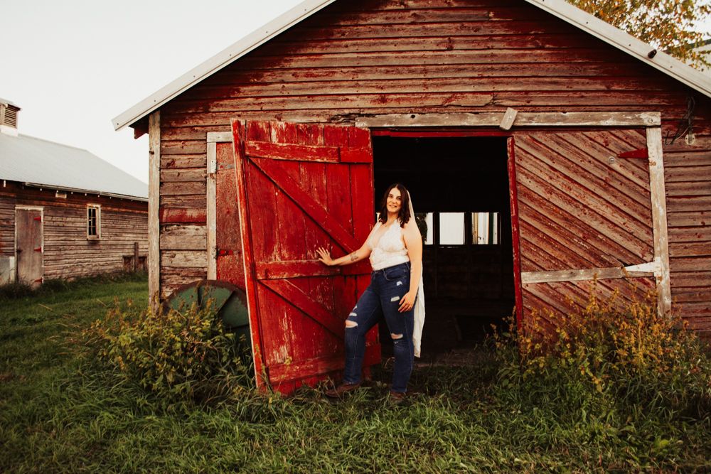 Figure in casual attire posing by red barn door in rustic countryside setting.