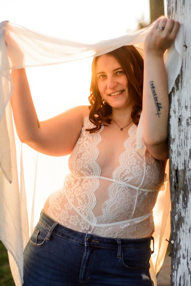 A figure in white lace and jeans poses against weathered wood in warm sunset lighting.