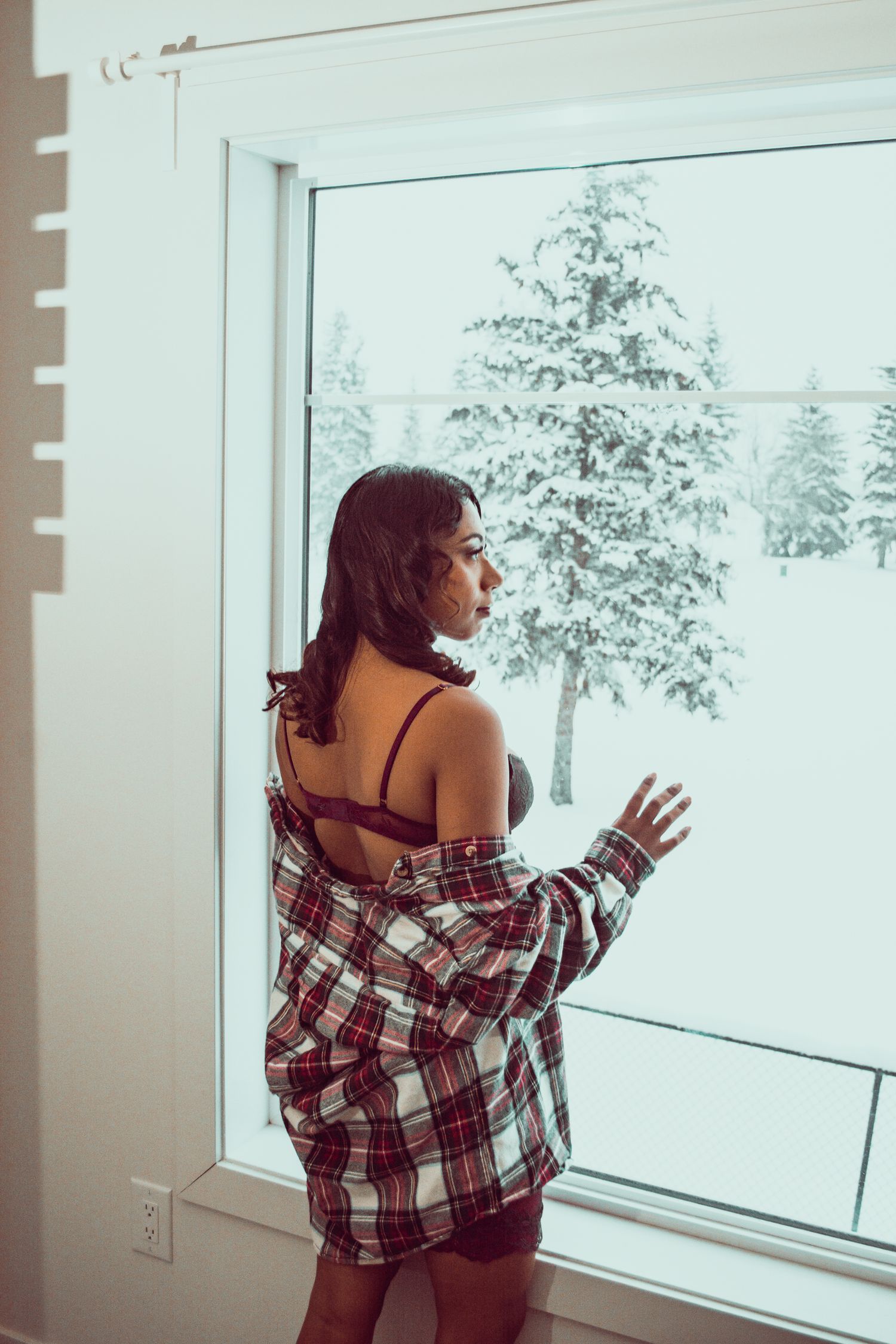 Person in plaid shirt looking through large window at snow-covered pine trees.
