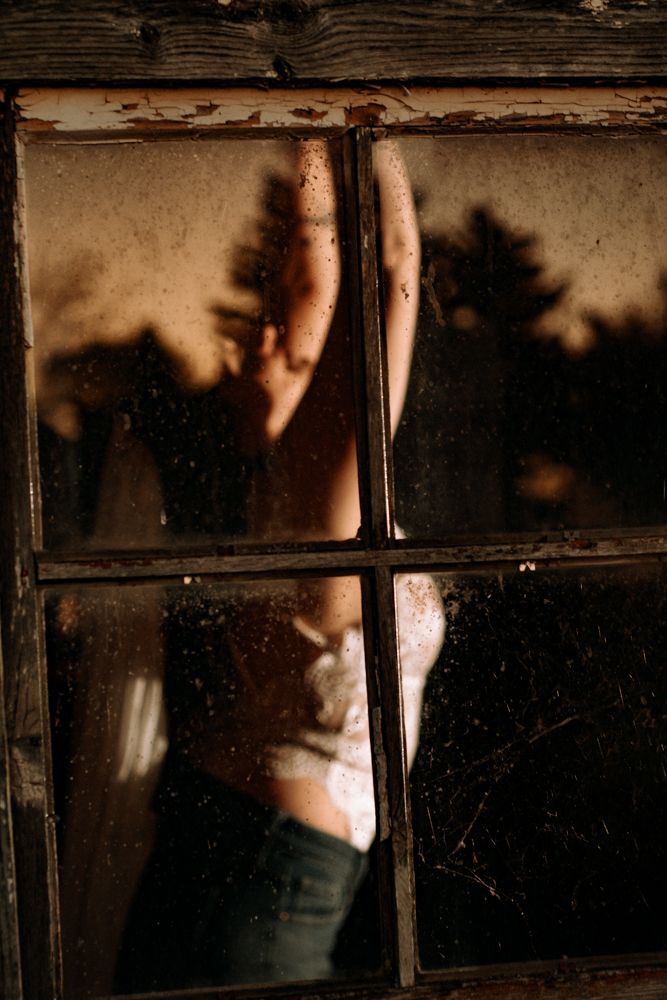 Moody portrait through weathered window panes with sunset trees in background.