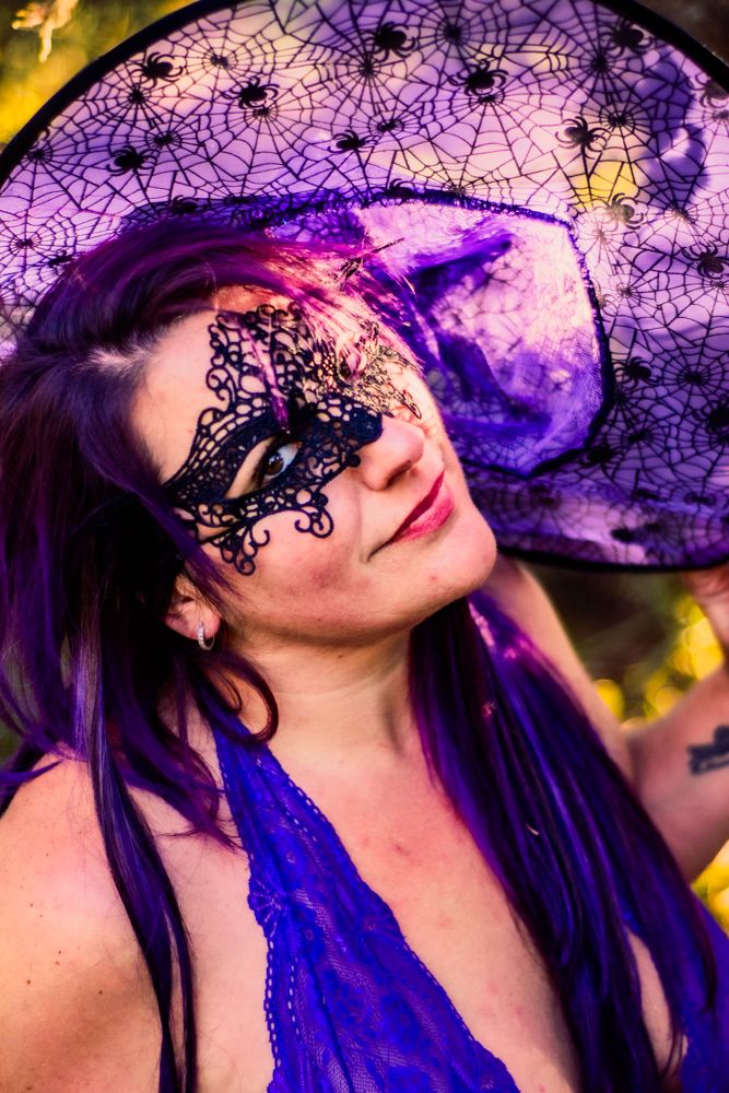 Intricate black lace mask and purple hat with spider web design against bokeh forest background.