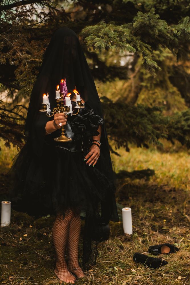 Mysterious figure in black dress and cape holds candelabra in forest setting with white candles on ground.