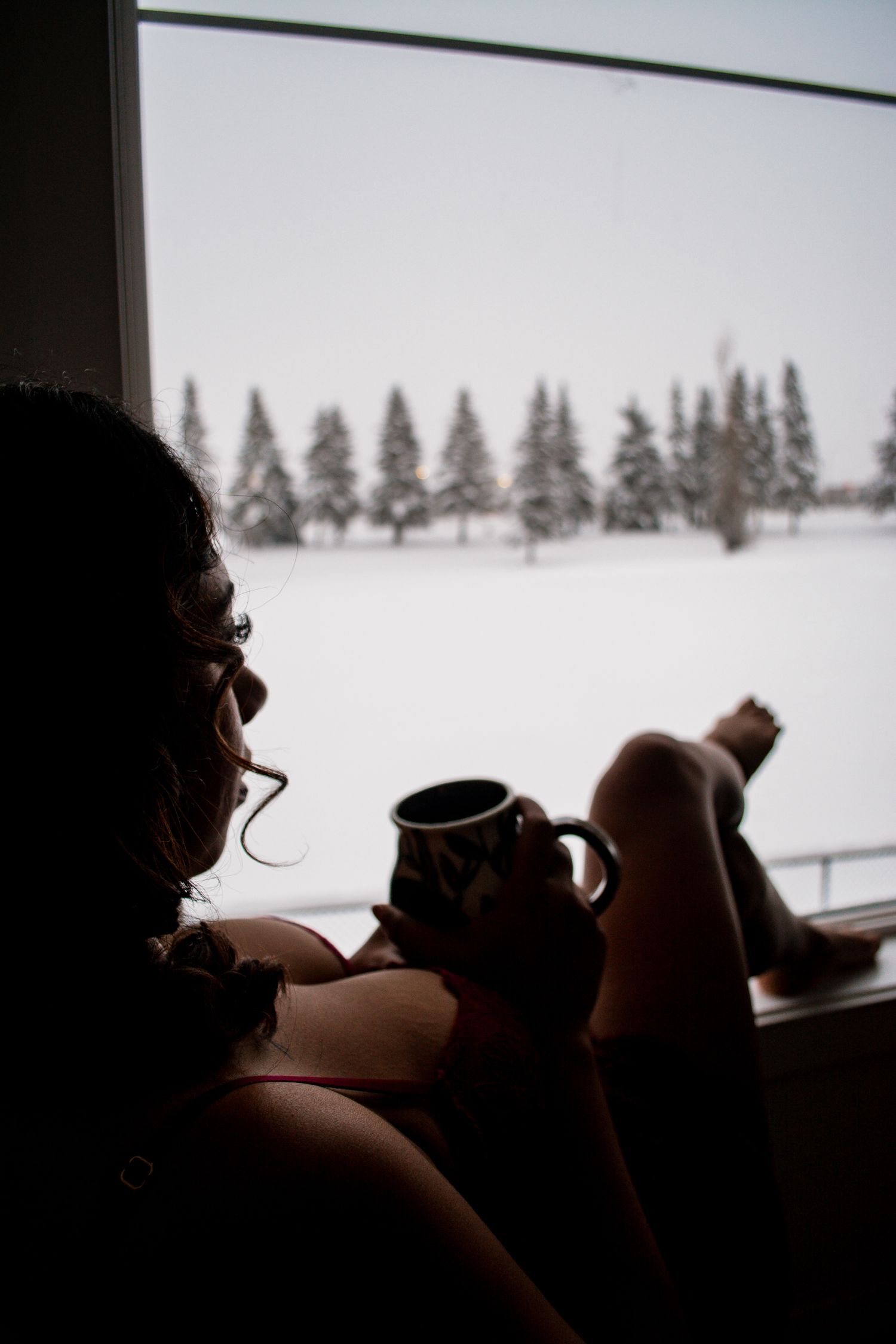 Silhouetted figure drinks coffee while gazing at snow-covered forest through window.