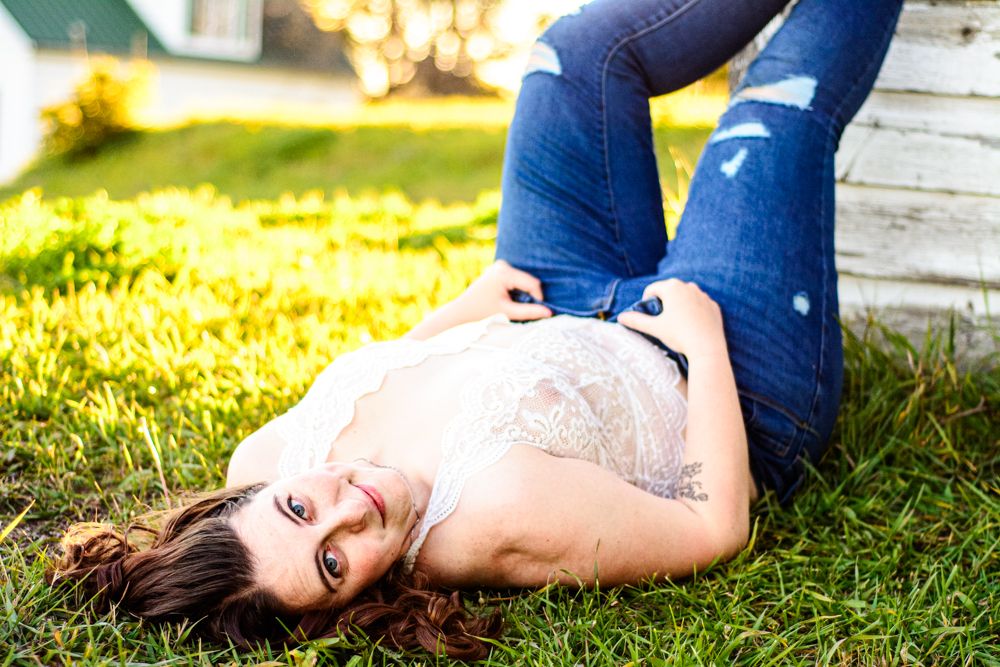 Model in white lace top and blue jeans relaxing on grass during golden hour photoshoot.