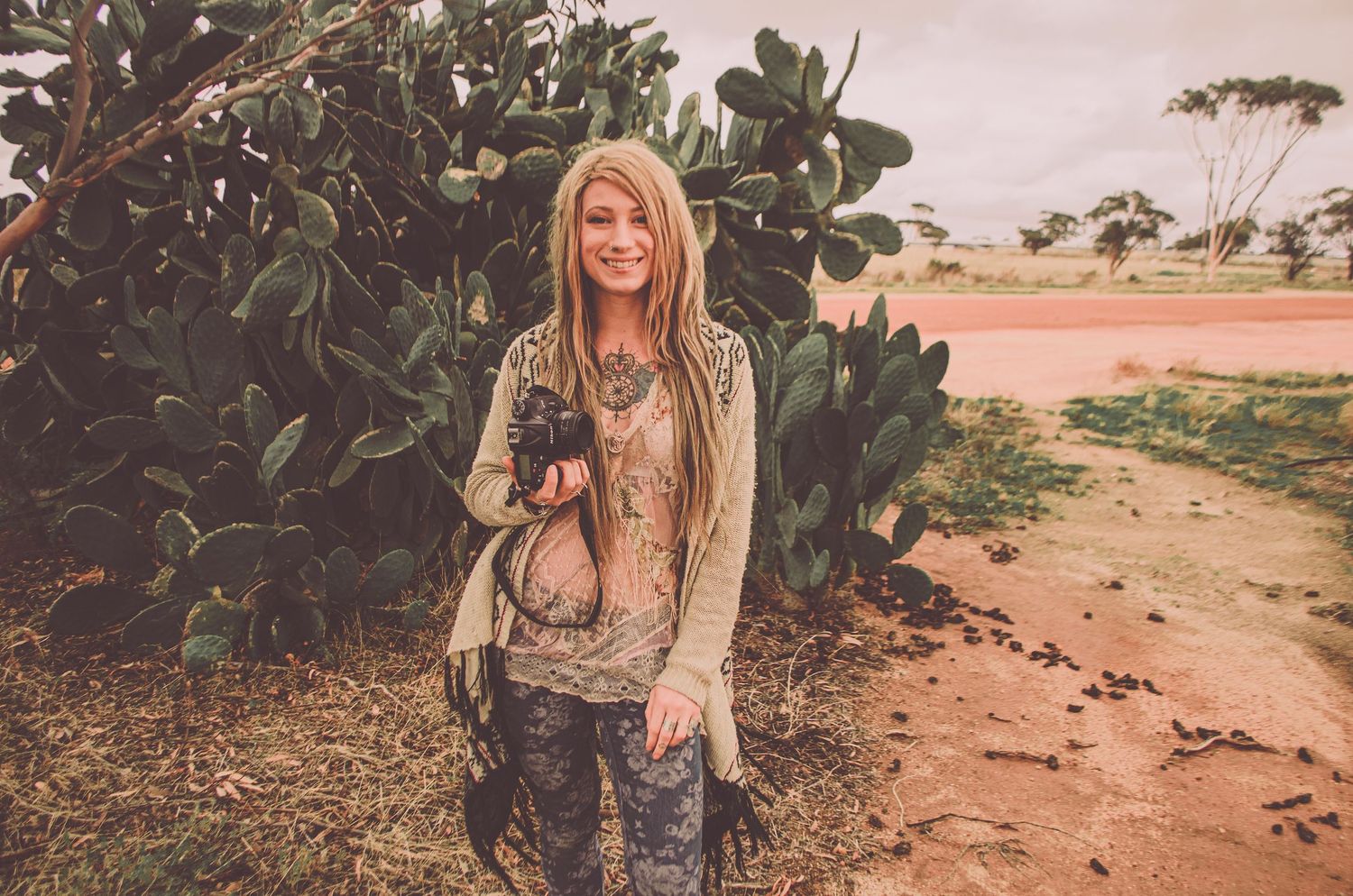 Young person in casual attire standing against a desert landscape with bushes at sunset.