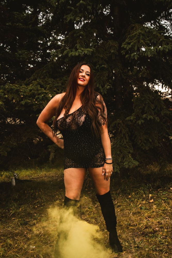 A woman in a black lace romper and knee-high boots poses outdoors with yellow smoke effects.
