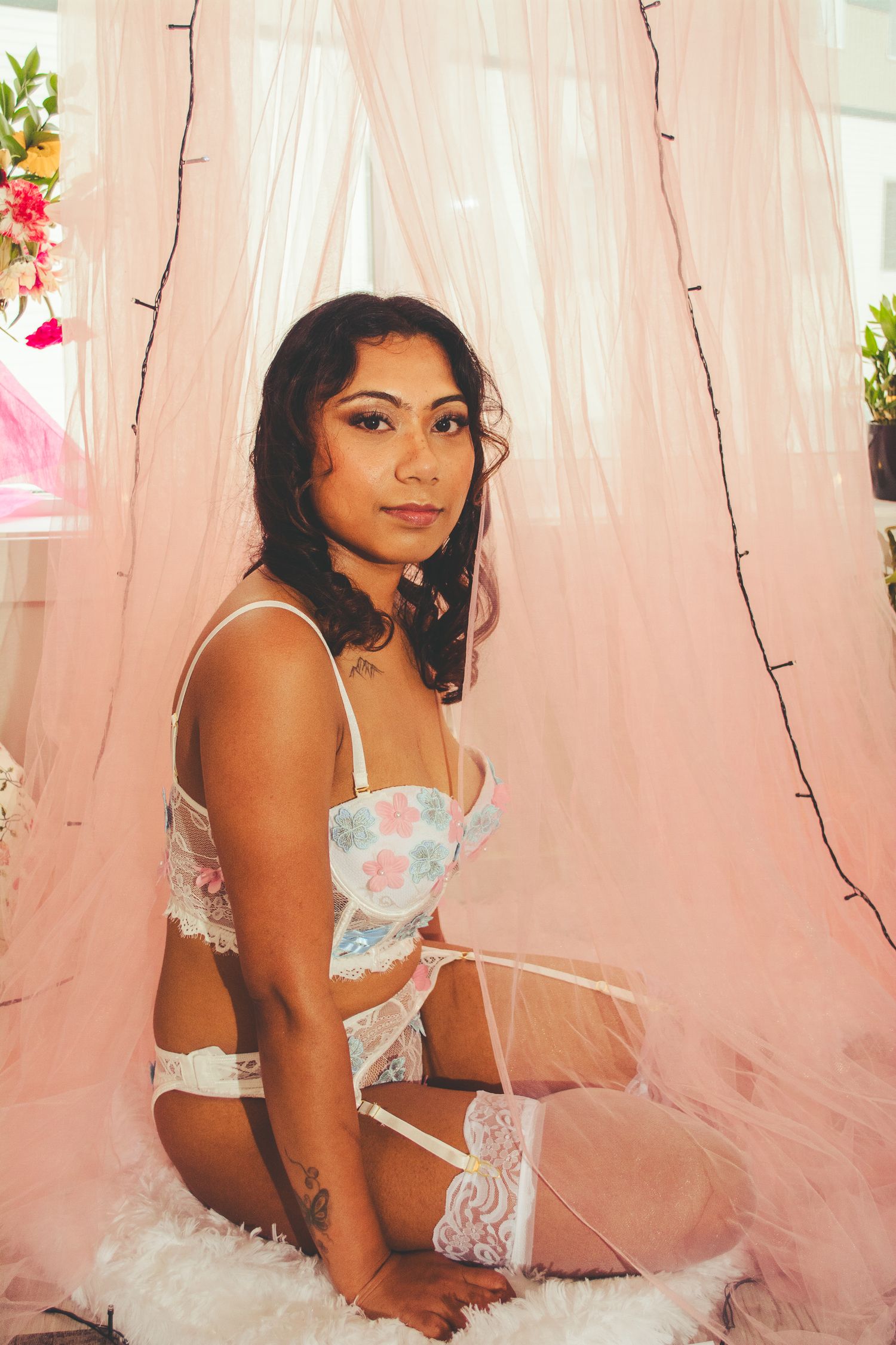 Boudoir photo shoot featuring soft pink curtains and romantic lighting with floral accents.