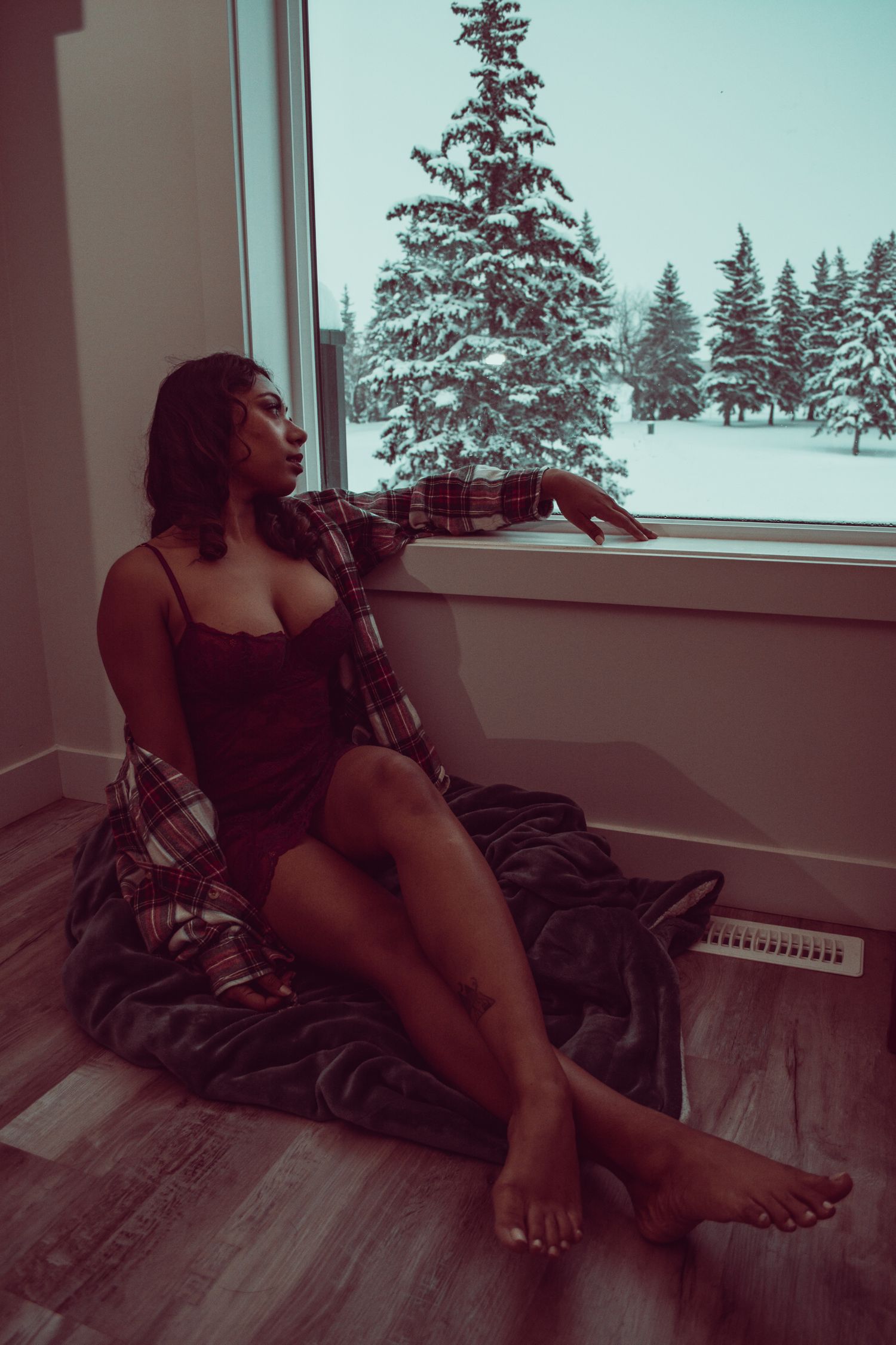 Woman in dark slip dress and plaid shirt gazes out window at snowy winter landscape.