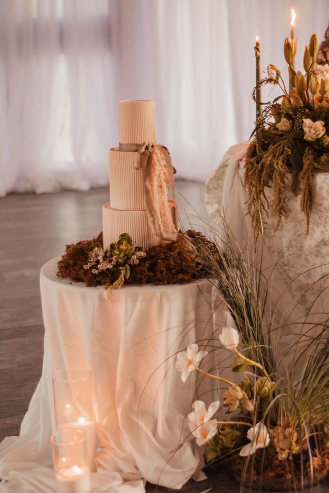 Elegant wedding cake display with dried flowers and soft lighting on white draped table.
