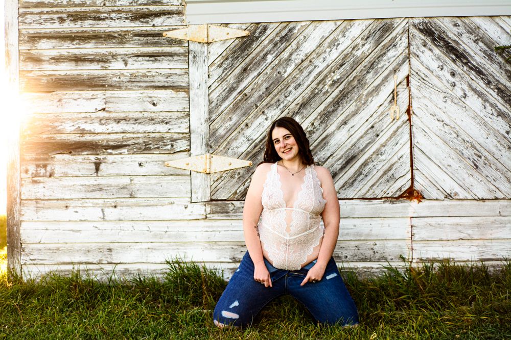 Person in lace top and jeans sitting against rustic white barn wall with sunlight.