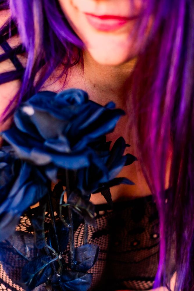 Close up of purple hair and red lips with a dark blue rose and black lace against a blurred background.